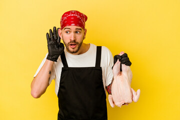 Young tattooed batcher caucasian man holding a chicken isolated on yellow background trying to listening a gossip.