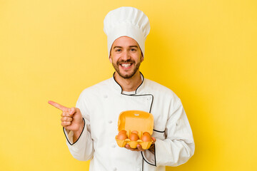 Young caucasian chef man holding eggs isolated on yellow background smiling and pointing aside, showing something at blank space.