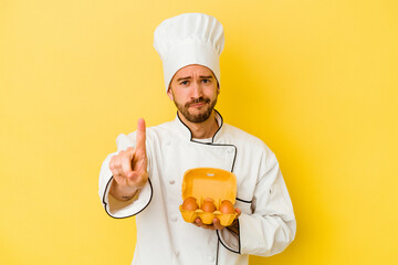Young caucasian chef man holding eggs isolated on yellow background showing number one with finger.