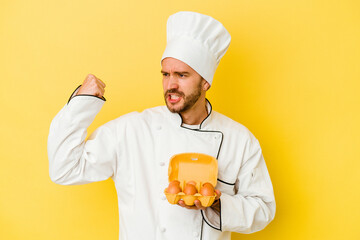 Young caucasian chef man holding eggs isolated on yellow background raising fist after a victory, winner concept.