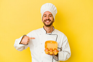 Young caucasian chef man holding eggs isolated on yellow background person pointing by hand to a shirt copy space, proud and confident