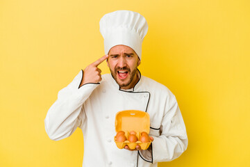 Young caucasian chef man holding eggs isolated on yellow background showing a disappointment gesture with forefinger.
