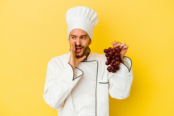 Young caucasian chef man holding grapes isolated on yellow background is saying a secret hot braking news and looking aside