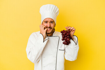 Young caucasian chef man holding grapes isolated on yellow background biting fingernails, nervous and very anxious.