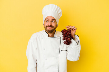 Young caucasian chef man holding grapes isolated on yellow background dreaming of achieving goals and purposes