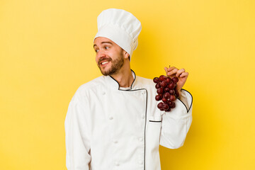 Young caucasian chef man holding grapes isolated on yellow background looks aside smiling, cheerful and pleasant.