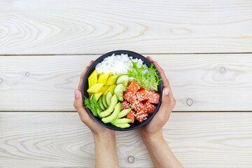 Poke a bowl of salmon, rice, avocado, cucumber, mango, herbs in a woman's hands. Hawaiian ahi, a...