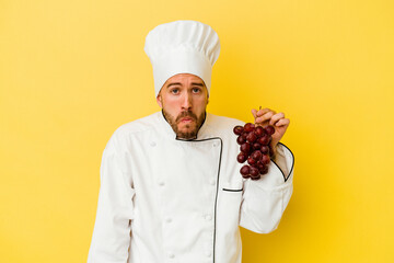 Young caucasian chef man holding grapes isolated on yellow background shrugs shoulders and open eyes confused.