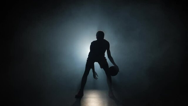 Silhouette of basketball player dribbles ball practicing skills in a dark studio with smoke and backlight. Professional player trains before basketball competition. Slow motion ready 59.94fps.