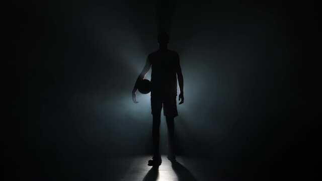 Out of darkness and smoke, silhouette of guy walking straight into the camera with a basket ball in his hands looms. Basketball player posing in studio with back light. Slow motion ready 59.94fps.
