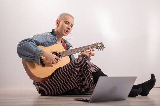 Young hipster woman sitting on the floor at home and watching guitar tutorials on laptop. Distance learning music in quarantine