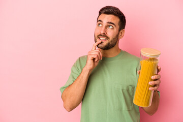 Fototapeta premium Young caucasian man holding pasta jar isolated on pink background relaxed thinking about something looking at a copy space.