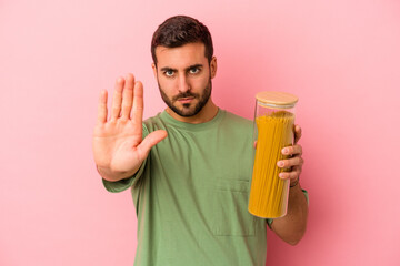 Young caucasian man holding pasta jar isolated on pink background standing with outstretched hand showing stop sign, preventing you.