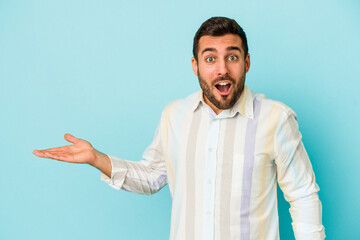 Young caucasian man isolated on blue background impressed holding copy space on palm.