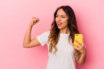 Young mexican woman holding a mobile phone isolated on pink background raising fist after a victory, winner concept.