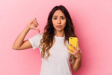 Young mexican woman holding a mobile phone isolated on pink background showing a dislike gesture,...