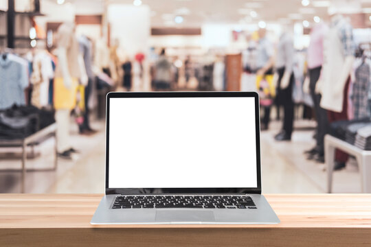 Empty screen laptop on a wooden table with abstract blurred background of interior clothing store at Shopping Mall