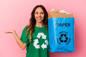 Young mexican woman recycling cardboard isolated on pink background showing a copy space on a palm and holding another hand on waist.