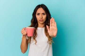 Young mexican woman holding a mug isolated on blue background standing with outstretched hand showing stop sign, preventing you.