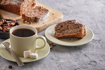 Delicious and flavorful banana cake with nuts and chocolate on a wooden board with a cup of coffee on a gray background