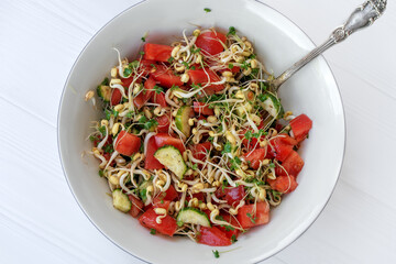 Salad with vegetables and microgreens, sprouts of cereals and broccoli, tomatoes, cucumbers, oil, salt, pepper in white bowl with metal spoon, on light background. Healthy energetic meal. Copy space.