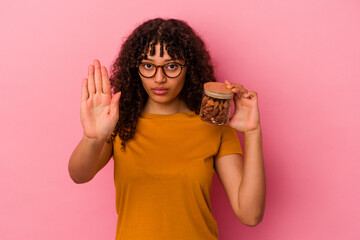 Young mixed race woman holding an almond jar isolated on pink background standing with outstretched hand showing stop sign, preventing you.