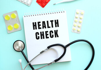 The text HEALTH CHECK is written on a white notepad that lies next to the stethoscope and pills on a blue background.
