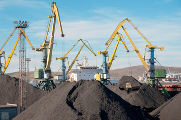 Fototapeta na wymiar Heaps of coal in the Murmansk Commercial Sea Port. Loading of coal by buckets (grabs) of portal cranes into the holds of a dry cargo ship in the seaport 