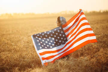 Female farmer stands with the American flag in a ripe wheat field against the backdrop of a beautiful sunset. The flag flies in the wind. Summer landscape.