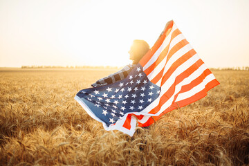 Female farmer holds a waving American flag against the backdrop of the sunset. Woman with a flag in a wheat field in summer. Independence Day. 4th of July.
