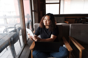 Smiling Asian young businesswoman is holding tablet for online shopping in social media at workplace in modern office or relaxing at coffee shop. Lifstyle of people with technology concept.