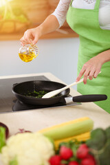 An attractive young dark-haired woman frying meal at worm pan by new keto recipe while standing and smiling in sunny kitchen. Cooking and householding concepts