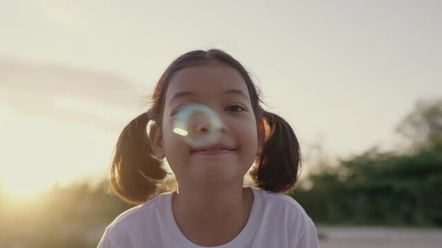 Happy Asian little girl playing to camera  in sunlight or sunset, Portrait of child smile enjoy playing soap bubbles and cheeky expression, Close up of face woman