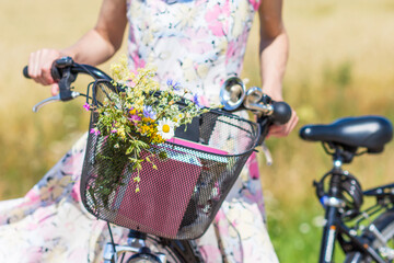 Wild flowers bouquet in basket of bicycle with woman in flowery dress in background, bike with...