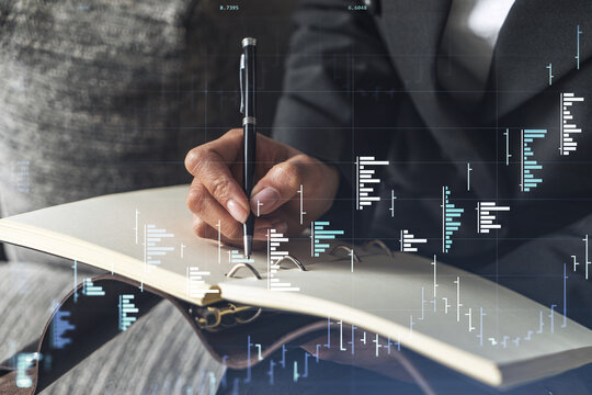 A woman trader in formal wear writing down some ideas to research stock market to proceed right investment solutions. Wealth management concept. Hologram Forex chart over close up shot.