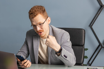 A businessman is sitting in a chair in the office. he holds the phone in one hand with an expression of delight, the other hand clenched into a fist