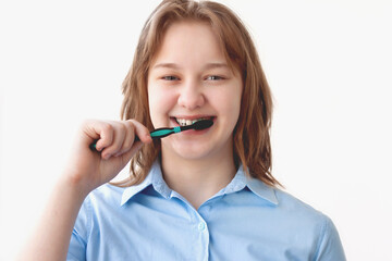 A girl in a blue shirt with blonde hair brushes her teeth and smiles against a light background, looking at the camera