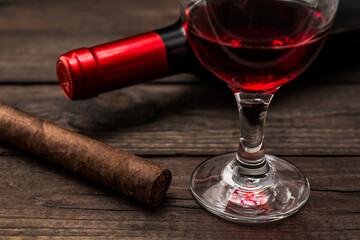 Bottle of red wine with a glass of red wine and cuban cigar on an old wooden table. Close up view, focus on the cuban cigar