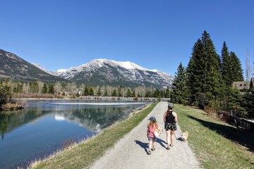 A mother and daughter going for a walk with their dog along the Bow River trails in Canmore, Alberta,  Canada on a beautiful sunny summer day. - obrazy, fototapety, plakaty