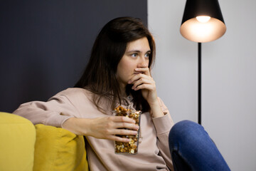 Attractive long-haired brunette girl sitting on Graphical background living room under spot lamp. pretty casual wearing lady hold cup of popcorn, relaxing at home, watching TV. Lazy Weekend concept