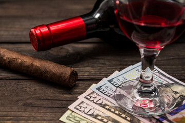 Bottle of red wine with a glass of red wine and cuban cigar with money on an old wooden table. Close up view, focus on the cuban cigar