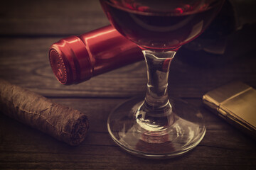 Bottle of red wine with a glass of red wine and cuban cigar with golden lighter on an old wooden table. Close up view, focus on the cuban cigar