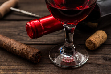 Bottle of red wine with a glass of red wine and cuban cigar with corkscrew on an old wooden table. Close up view, focus on the cuban cigar