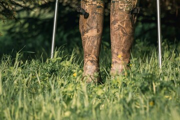 Legs in camouflage boots and trekking poles in the green grass among the trees. Hiking concept