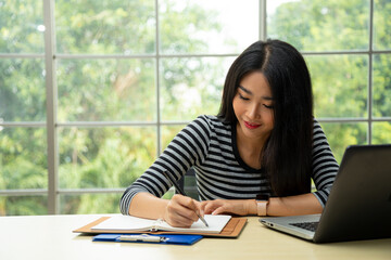 Happy young Asian woman smiling writing memo on notebook taking data from laptop in living room with copy space. teenage work from home concept