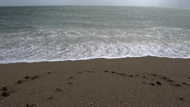 Wide angle slow motion footage of waves gently coming into shore and back out again. Camera pans up towards horizon line and blue sky. Calm tranquil footage of UK coast.