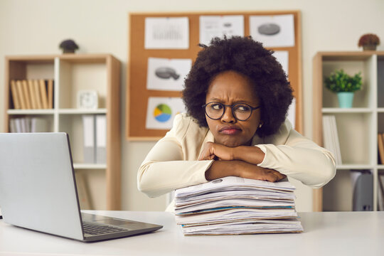 Lazy displeased fatigue young frowning african american woman worker employee keeping chin on paper stack folder pile sitting at table in office. Work overtime load, lots of paperwork concept