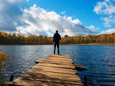 A young man stands on a wooden bridge near a forest lake. Enjoy the autumn landscape and nature . Freedom and travel