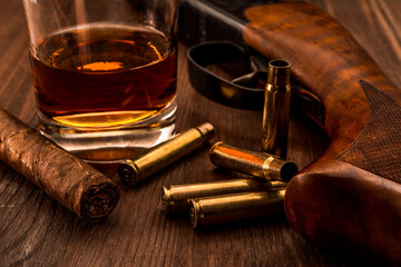 Empty shells and the rifle with glass of whiskey and cuban cigar on a wooden table. Focus on the cuban cigar