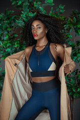 Portrait of a sensual young black woman with long curly black hair, beautiful makeup posing by herself in a studio with a green leave background wearing a blue fitness outfit with a brown trench coat.